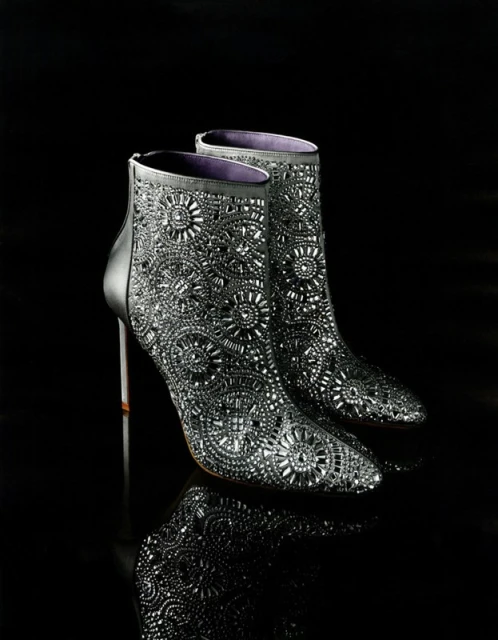 Elevated view of a pair of black, sparkling, ornate ankle boots with stiletto heels are displayed on a reflective black surface.