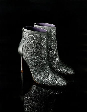 Elevated view of a pair of black, sparkling, ornate ankle boots with stiletto heels are displayed on a reflective black surface.