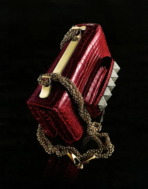 Elegant red crocodile-patterned handbag with golden chain and pyramidal studs, set against a stark black backdrop with subtle reflections.