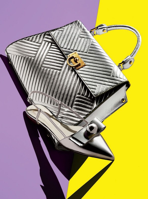 Still life studio shot of a silver metallic handbag and matching heeled slingback shoe on a split yellow and purple background.