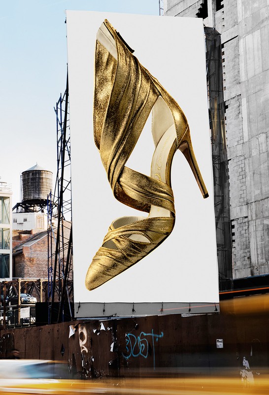 Billboard advertisement for a gold high-heeled shoe with a blurred urban street scene, buildings, and a water tower in the background.