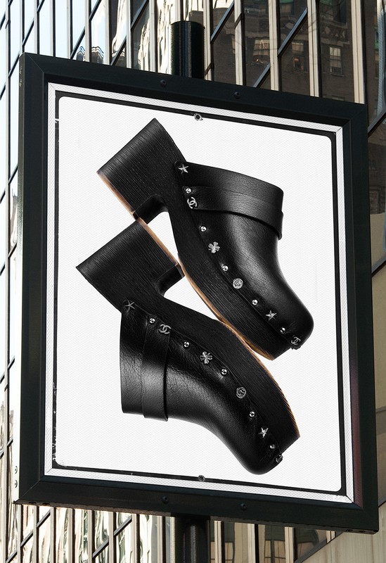 Fashion advertisement featuring two black leather clogs with studded details against a blurred building backdrop.