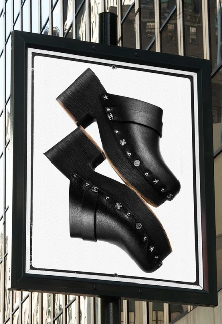 Fashion advertisement featuring two black leather clogs with studded details against a blurred building backdrop.