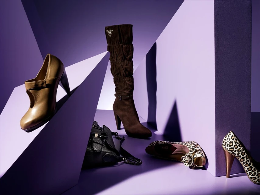 Fashion still life showcasing a brown Prada boot, beige heeled pump, leopard print heels, and a black purse against a geometric purple backdrop.