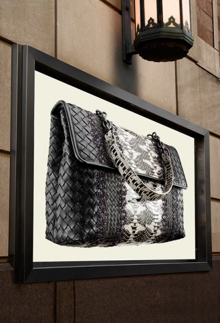 Fashion handbag display with black woven leather and white snake skin design mounted on an exterior building wall next to a light fixture.