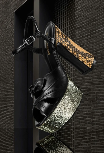 Elevated studio shot showcases a platform sandal with snakeskin heel, glittery platform, and black leather straps against a dark brick backdrop.