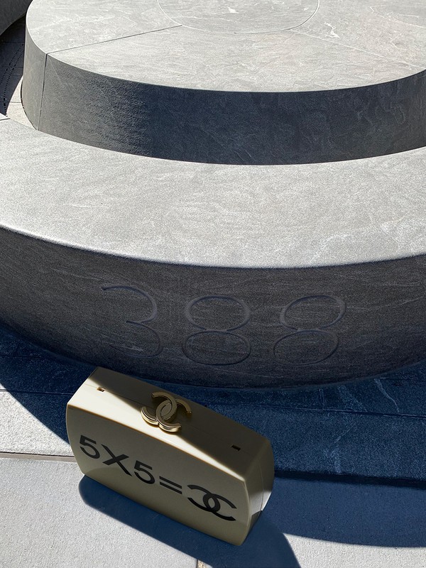 Beige clutch with gold Chanel logo on a concrete surface against a tiered gray platform with inscribed numbers; luxury fashion accessory.