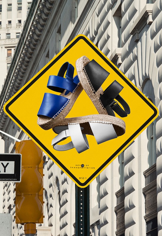 Fashion-forward product shot of blue, white, and black strappy sandals arranged atop a yellow street sign, set against a building exterior.
