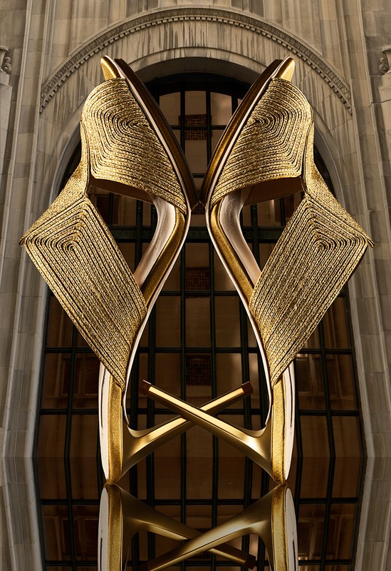 Gold platform shoes on a reflective surface, displayed against an architectural backdrop with an arched window and stone facade.