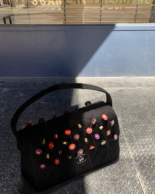 Designer black handbag with colorful button embellishments and logo detail on a textured surface with a building reflection in the background.
