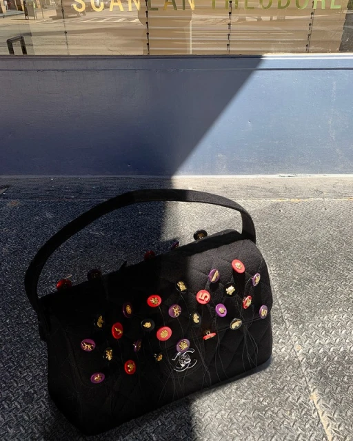Designer black handbag with colorful button embellishments and logo detail on a textured surface with a building reflection in the background.