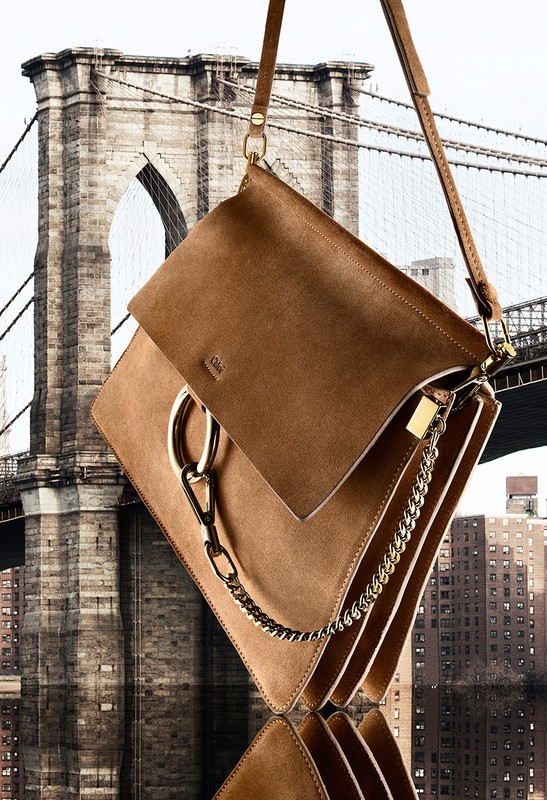 Brown leather shoulder bag with a chain strap against a background featuring a cityscape and bridge architecture.