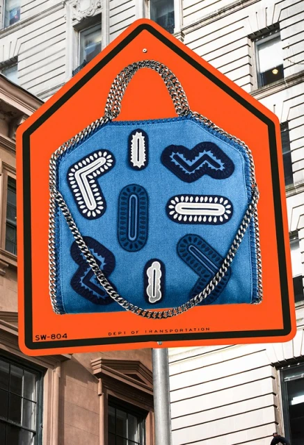 Denim shoulder bag with chain straps mounted on orange road sign in urban setting with building and sky in the background.