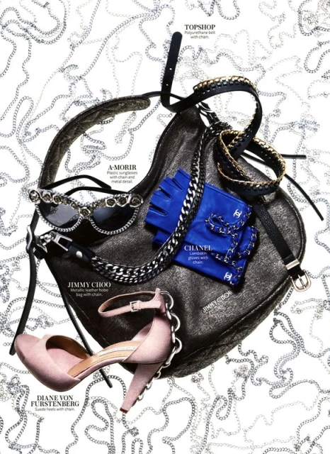 Fashion flat lay featuring a black purse, pink heels, blue gloves, sunglasses, and a black belt, all accented with silver chains. 
