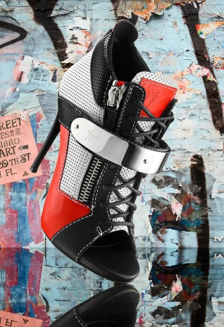 Elevated view of a black, white, and red high-heeled shoe against a graffiti background. The shoe has laces, zippers, and a shiny metal strap.