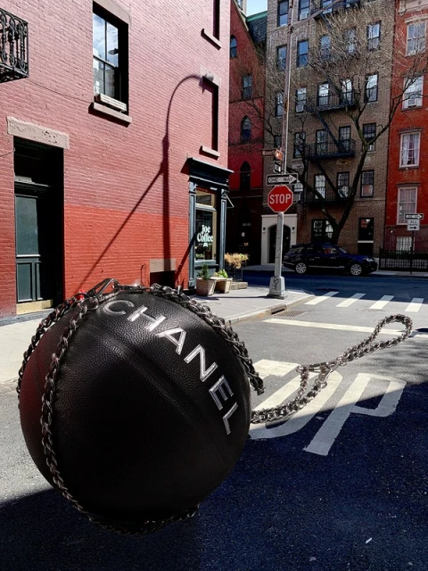 Chanel ball-shaped handbag with chain strap on asphalt street in an urban setting with red brick buildings and storefronts in the background.