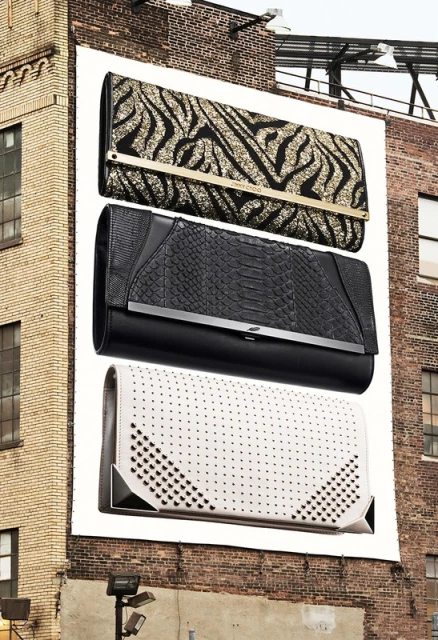 Outdoor billboard advertisement features three fashion clutches with patterns and metallic accents, mounted to the side of a brick building.