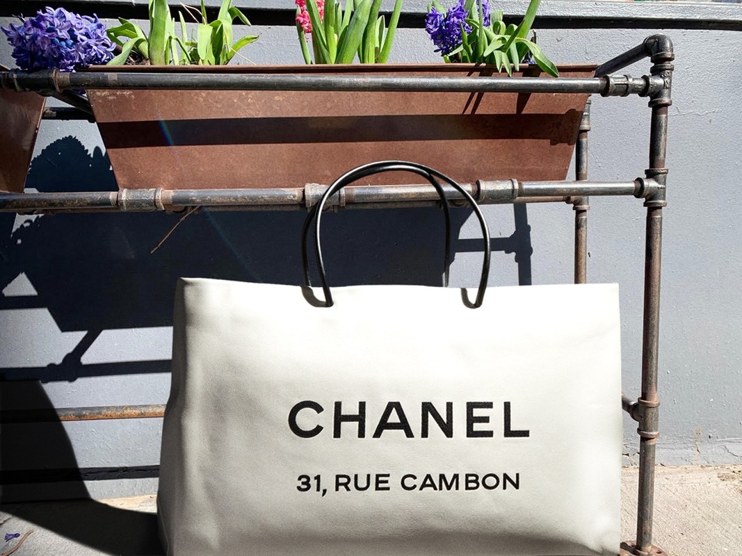 Ivory Chanel shopping bag with black handles and logo displayed against a gray wall and planter in an outdoor setting.