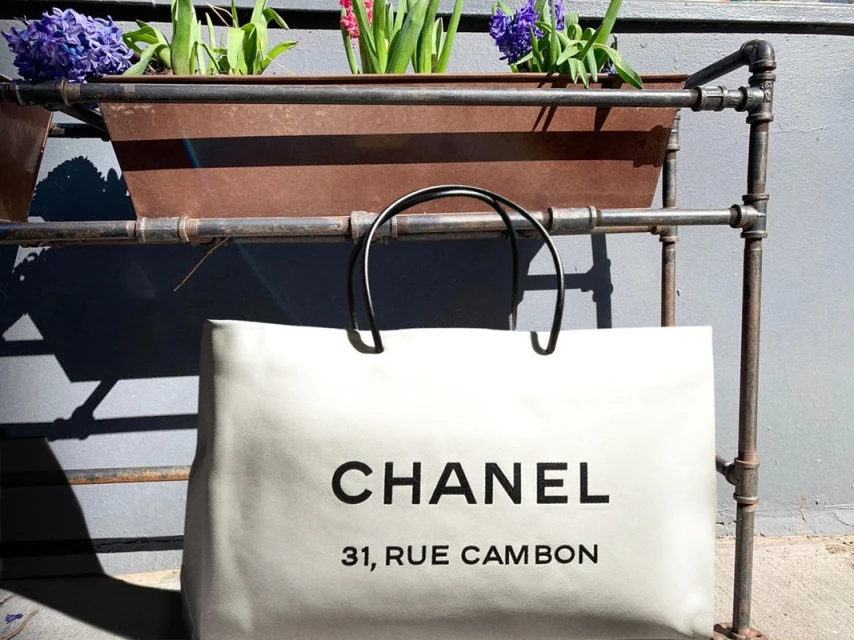 Ivory Chanel shopping bag with black handles and logo displayed against a gray wall and planter in an outdoor setting. 