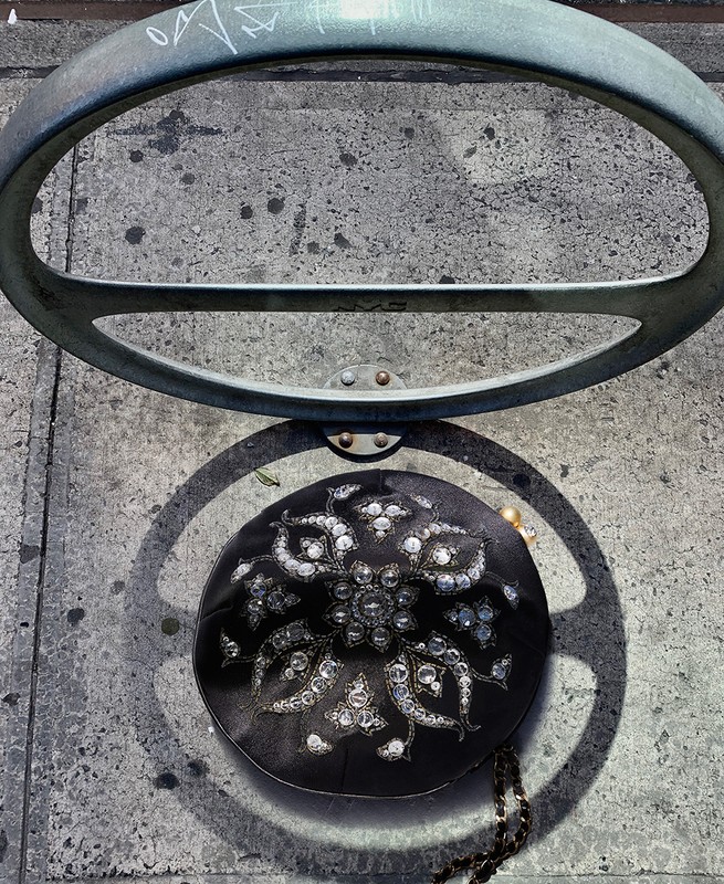 Overhead shot of a black clutch purse with decorative jewels placed on a concrete ground next to a metal bike rack.