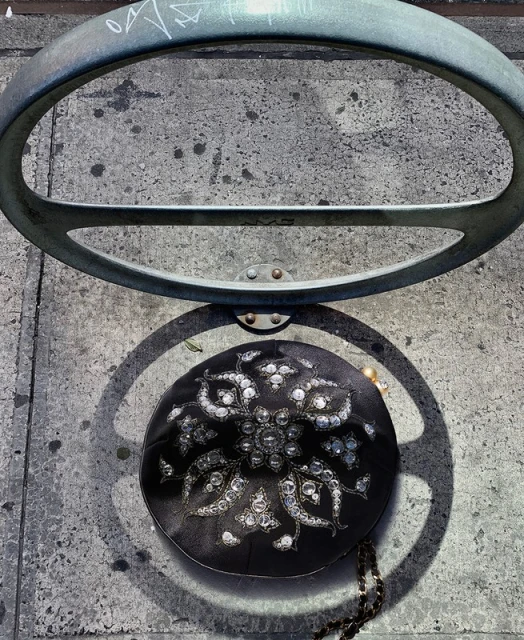 Overhead shot of a black clutch purse with decorative jewels placed on a concrete ground next to a metal bike rack.