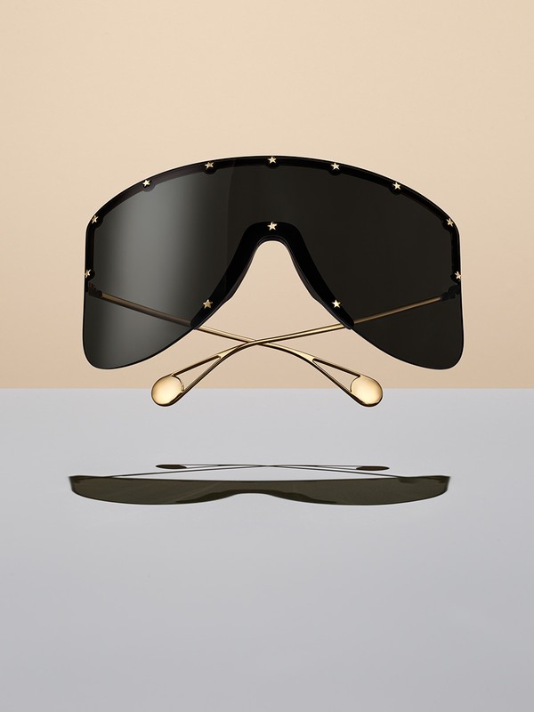 Oversized black shield sunglasses with gold frames and star details hover against a split beige and light grey backdrop. Product shot.