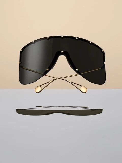 Oversized black shield sunglasses with gold frames and star details hover against a split beige and light grey backdrop. Product shot.