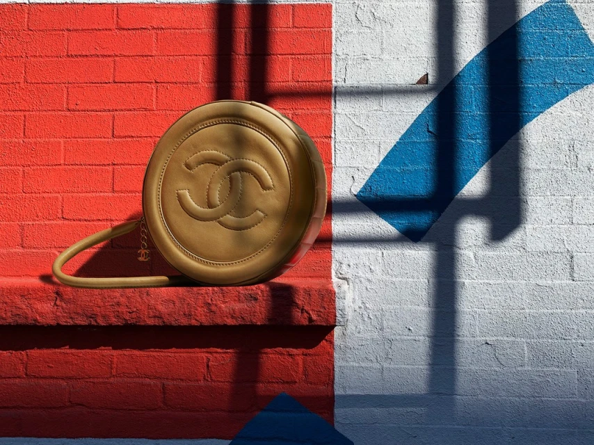 Round tan purse with the Chanel logo rests on a red brick ledge against a white brick wall with a blue stripe and shadows.