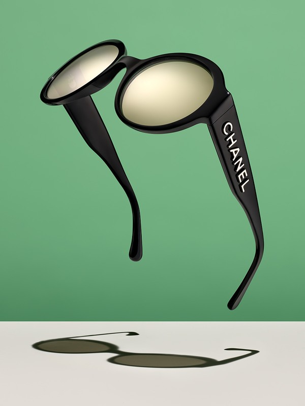 Studio shot of black Chanel sunglasses floating in front of a split green and off-white backdrop with visible shadow.