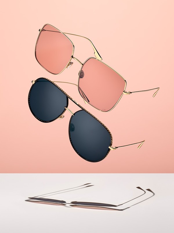 Still life studio shot of two pairs of gold-framed sunglasses with different colored lenses, casting shadows on a white surface.
