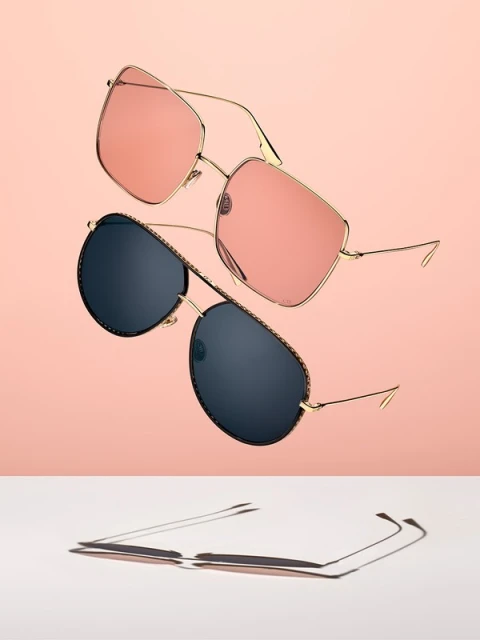 Still life studio shot of two pairs of gold-framed sunglasses with different colored lenses, casting shadows on a white surface.