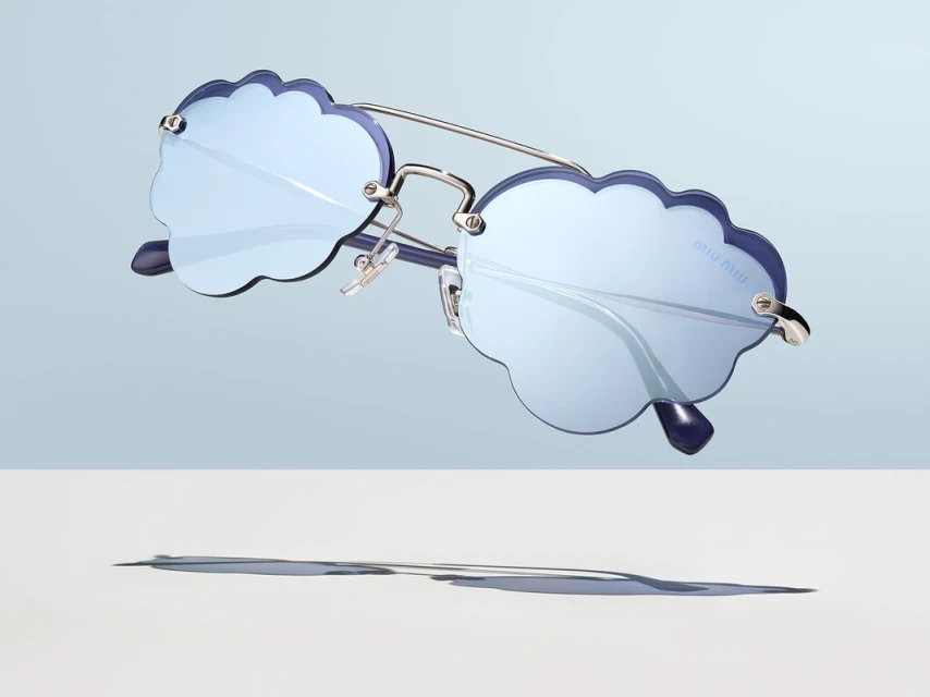 Cloud-shaped Miu Miu sunglasses in light blue with silver metal frames and dark blue rims, casting a shadow on white surface.
