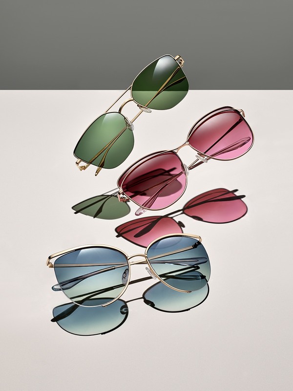 Still life studio shot featuring three pairs of gold-framed sunglasses with green, red, and blue gradient lenses, positioned on a gradient backdrop.