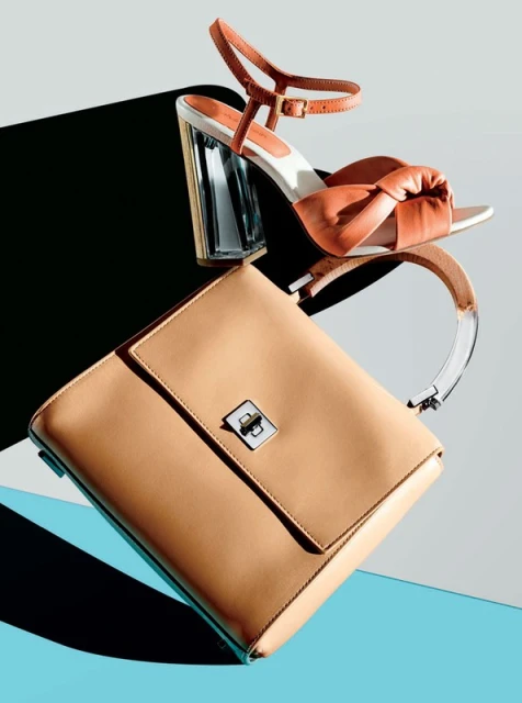 Tan leather handbag and coral colored heeled sandal on a turquoise and grey background, studio product photography.