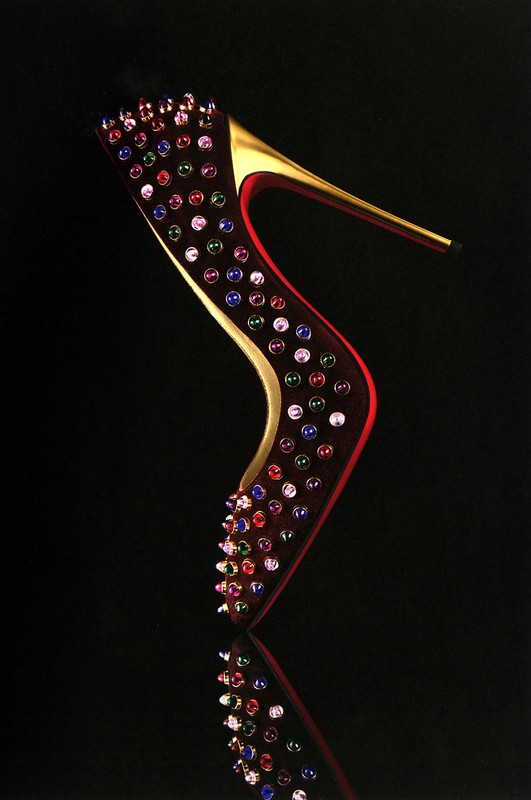 Elegant high-heeled shoe with golden heel and colorful gemstone embellishments, displayed against a sleek black backdrop with reflective surface.