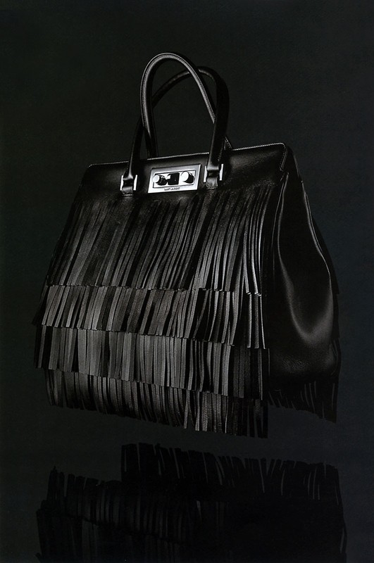 Sleek black leather handbag with fringe detail. Fashionable accessory with a glossy finish, displayed against a dark background with reflection.