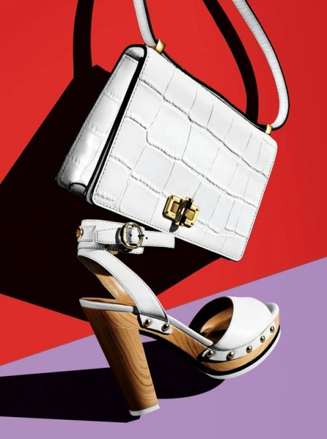 Fashion still life of a white crocodile-skin handbag and a white sandal with a wooden heel against a vibrant red and purple backdrop.