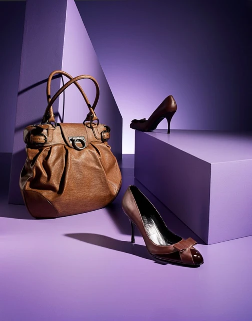 Studio shot of a brown leather handbag and brown leather high heel shoes arranged on a purple surface against a geometric background.