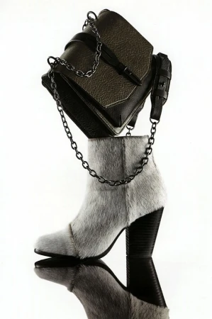 Fashion still life featuring a leather handbag with chain strap balanced on a furry ankle boot against a white background with reflection.