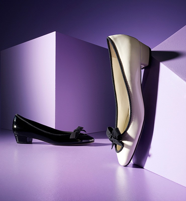 Elegant black and white patent leather heeled pumps with bow detail against a purple studio backdrop for fashion product shot.