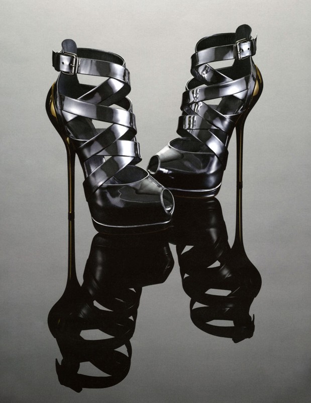 Pair of black strappy high heels with open toes, buckles, and stiletto heels reflecting on a glossy gray surface in a studio shot.