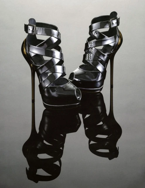 Pair of black strappy high heels with open toes, buckles, and stiletto heels reflecting on a glossy gray surface in a studio shot.