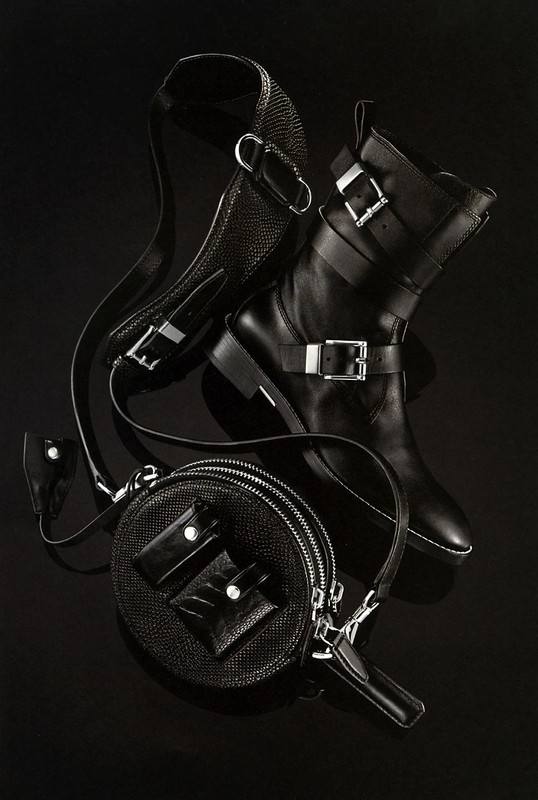 High-angle shot of a black leather ankle boot with silver buckle, a round crossbody bag, and a shoulder strap, all against a black background.