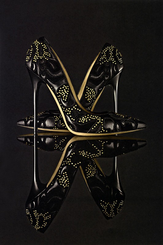 Pair of black, high-heeled shoes are mirrored against a dark background, showcasing an elegant design with decorative embellishments.
