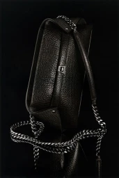 Close-up of a black textured leather shoulder bag with a chunky metal chain strap against a dark background.