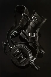 Black leather combat boot, round zippered pouch, and textured strap arranged on reflective black background.