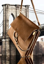 Tan suede shoulder bag with gold hardware suspended against a blurred backdrop of a suspension bridge.