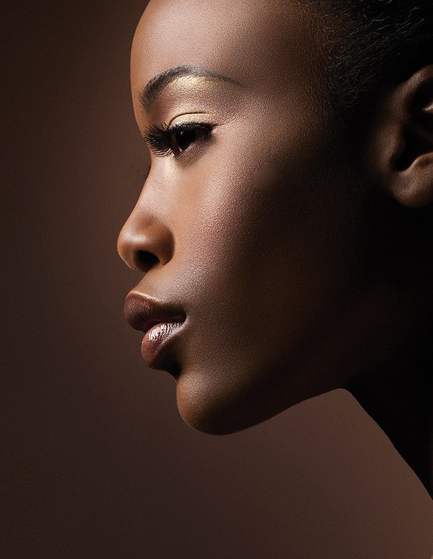 Close-up studio shot of a young, attractive Black woman with golden eyeshadow and glossy lips against a brown background.