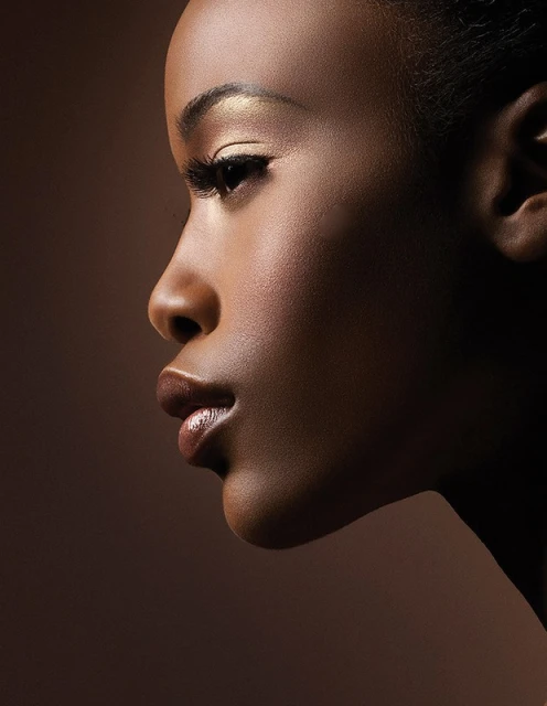 Close-up studio shot of a young, attractive Black woman with golden eyeshadow and glossy lips against a brown background.