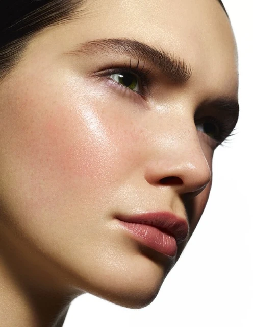 Close-up portrait of a woman with fresh, dewy makeup, focusing on her radiant skin, arched eyebrows, and subtle lip color against a white background.
